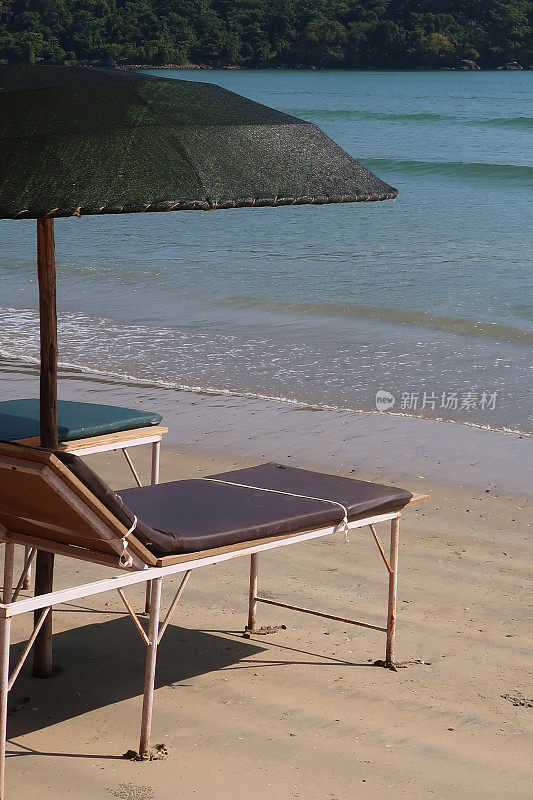 在热带沙滩上，木制太阳躺椅撑着阳伞，沙滩浴巾晒日光浴，在树荫下睡觉，木制太阳躺椅热带海滩假日海滨度假印度果阿/喀拉拉邦